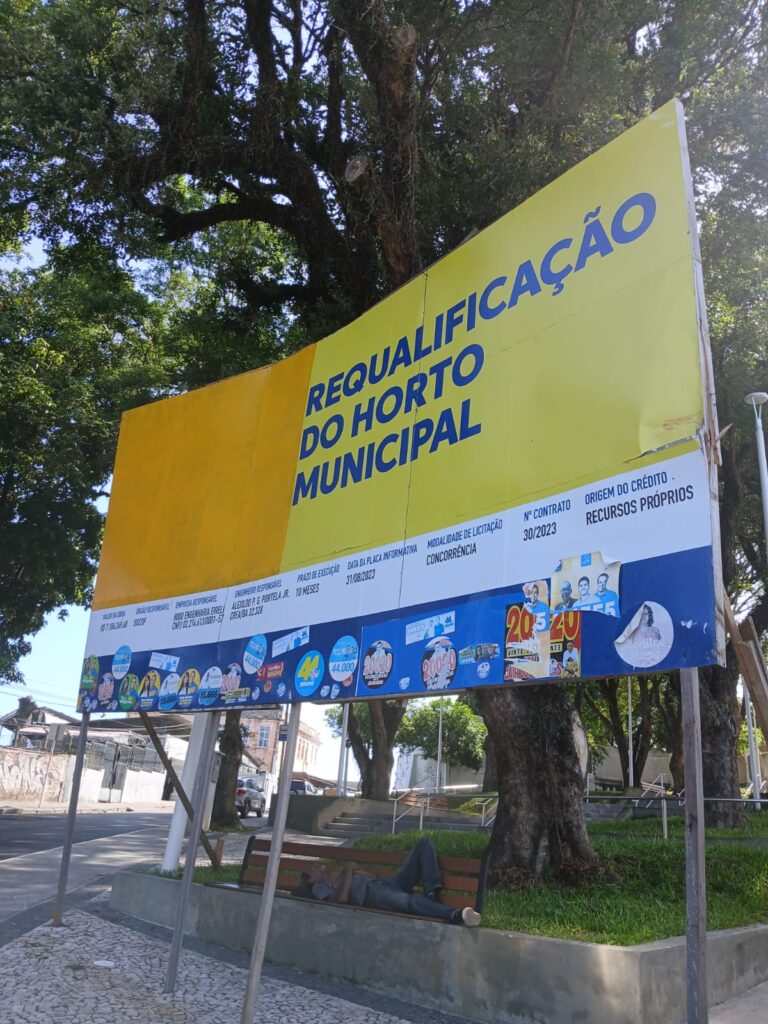 Imagem da placa de anúncio da requalificação do Horto Municipal no Bairro do Bonfim. A placa está nas cores amarelo e azul. Atrás há a presença de árvores e pavimentação.