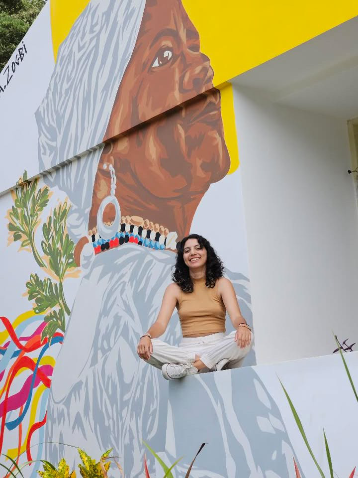 Na imagem há a figura de uma menina sentada a frente de uma mural bem colorido, com elementos representativos da cultura baiana.