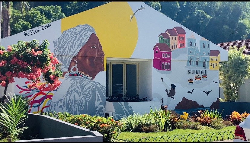 Na imagem há um grande mural na parede da Sumai/UFBA. Na obra há ilustrações de elementos da cultura baiana como a baiana de acarajé, o pelourinho e a roda de capoeira.