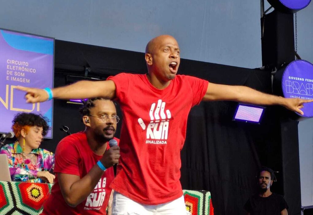 No centro da imagem está Elinilson Soares, homem negro, surdo, com uma expressão intensa e braços abertos, vestindo uma camiseta vermelha com a imagem de uma mão aberta, símbolo do coletivo Rua Sinalizada. Próximo a ele está Lucas Sol, homem negro sem deficiência e usando uma camisa vermelha semelhante a de Elinilson. Ambos estão em cima de um palco e atrás deles está uma pessoa sentada em uma mesa enquanto outra observa de longo, com um painel ao fundo. Na frente do palco, se destaca uma placa, novamente com o símbolo do coletivo.